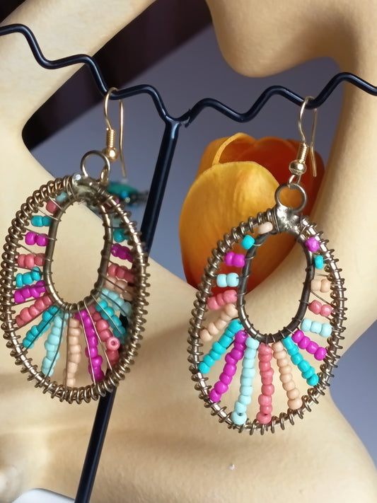 Paire de boucles d’oreilles pendantes de forme ovale, cerclées de métal torsadé et décorées de perles multicolores alternant les tons roses, bleus et beiges, présentées sur un support dédié.