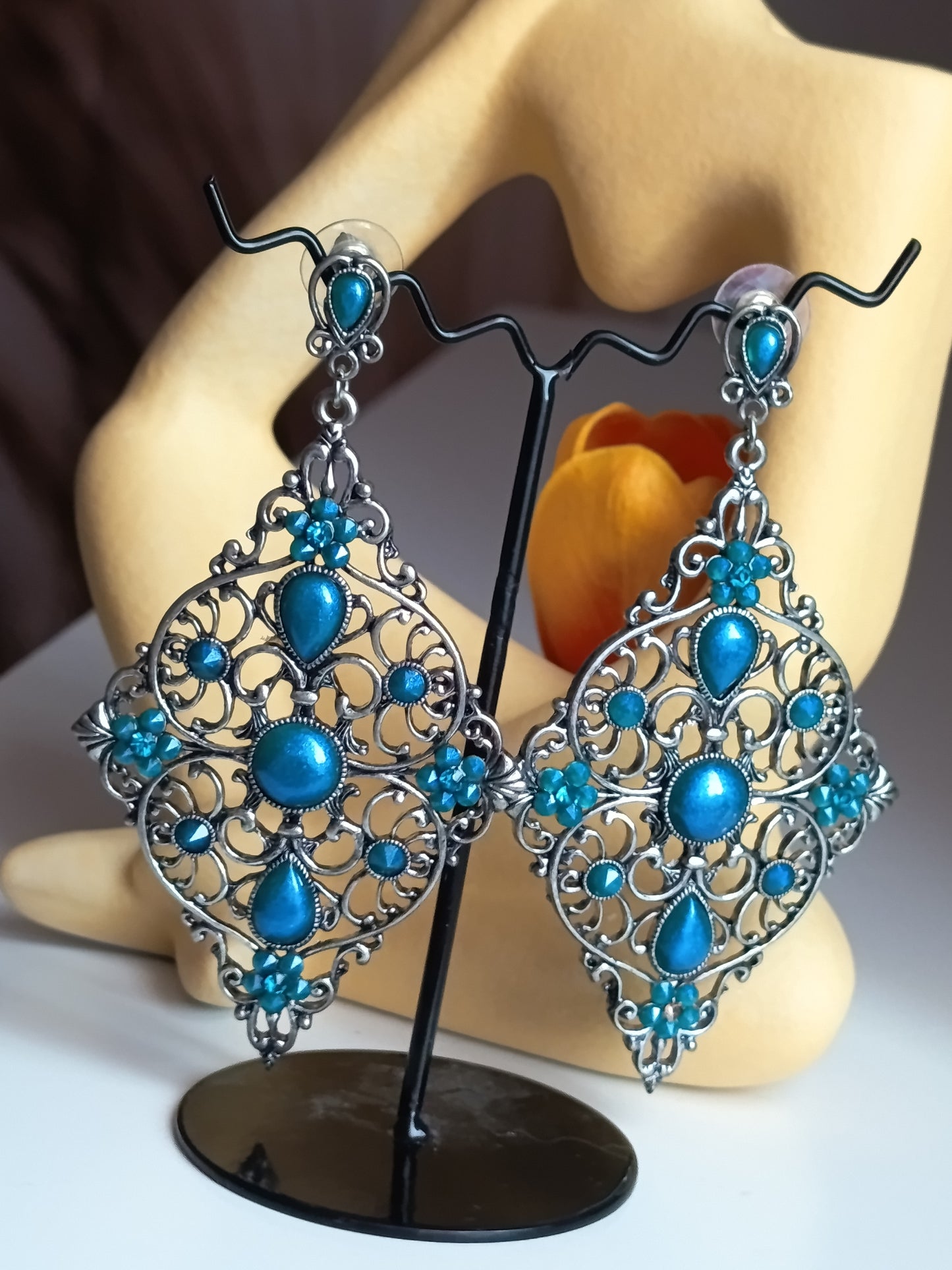 Paire de boucles d'oreilles pendantes de style bohème chic, en métal argenté avec des ornements complexes en filigrane et serties de multiples pierres et cabochons bleu turquoise éclatant. Elles sont présentées sur un support noir devant un mannequin de présentation et un élément.