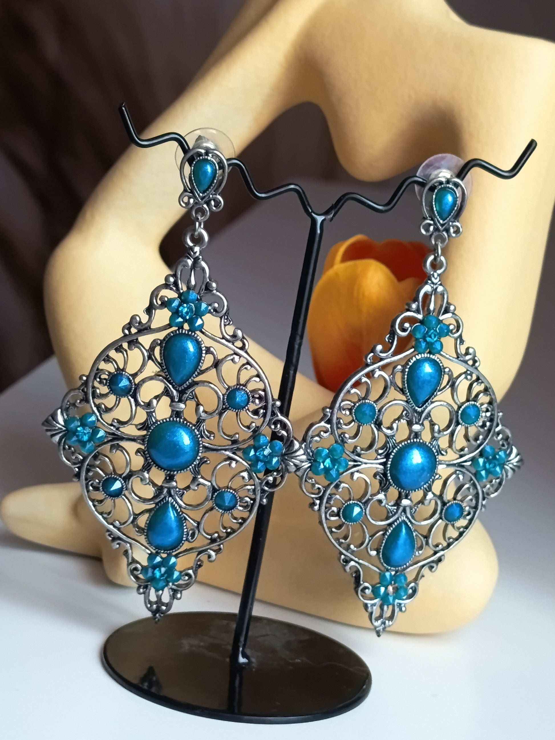 Paire de boucles d'oreilles pendantes de style bohème chic, en métal argenté avec des ornements complexes en filigrane et serties de multiples pierres et cabochons bleu turquoise éclatant. Elles sont présentées sur un support noir devant un mannequin de présentation et un élément.