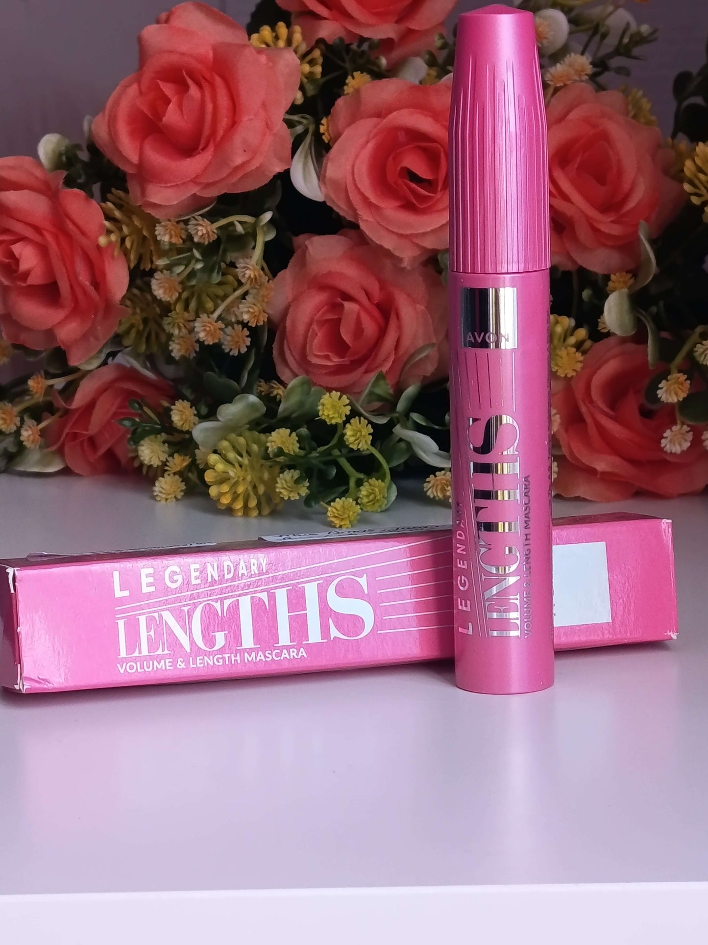 "Tube de mascara rose Legendary Lengths d'Avon posé devant sa boîte cartonnée rose et un bouquet de roses corail."