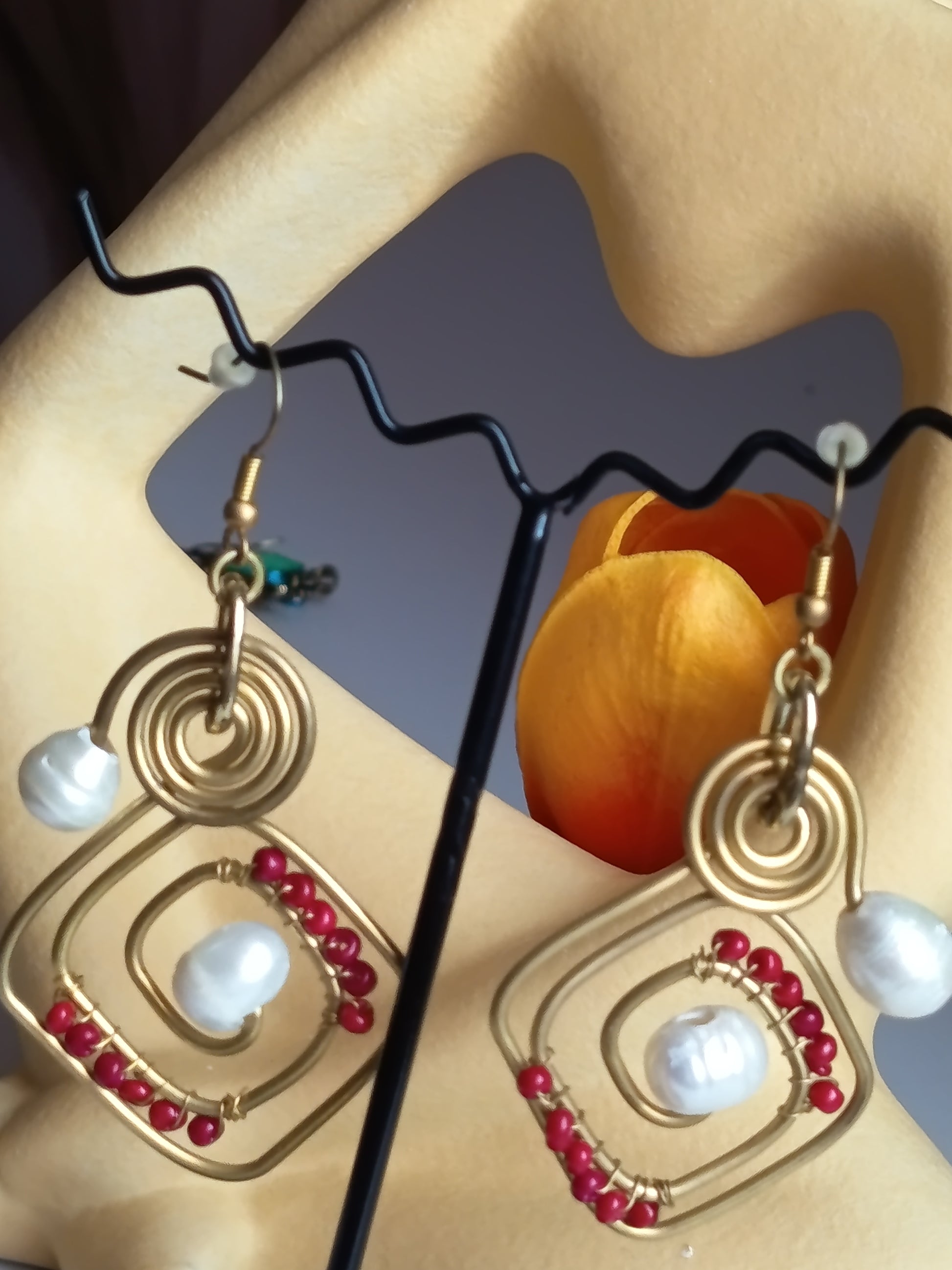 Boucles d'oreilles pendantes artisanales, motif géométrique en métal doré avec perles blanches et perles rouges, présentées sur un support ✨