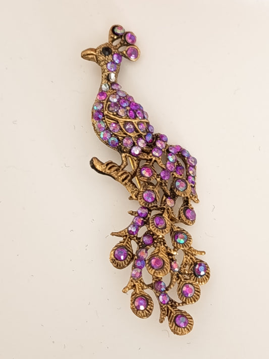 Broche en forme de paon dorée sertie de cristaux violets et strass aurore boréale sur fond blanc. 