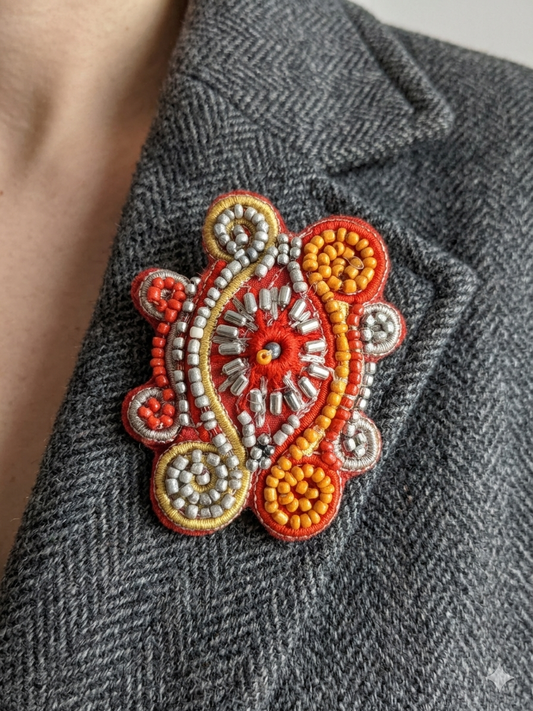 Broche artisanale brodée main avec perles de rocaille orange et argent, cannetille dorée sur fond rouge. Portée sur une veste habillée. 