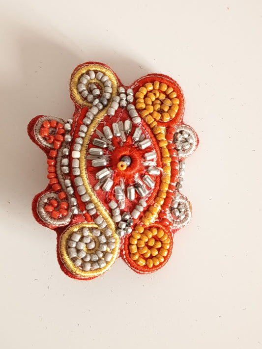 Broche artisanale brodée main avec perles de rocaille orange et argent, cannetille dorée sur fond rouge.