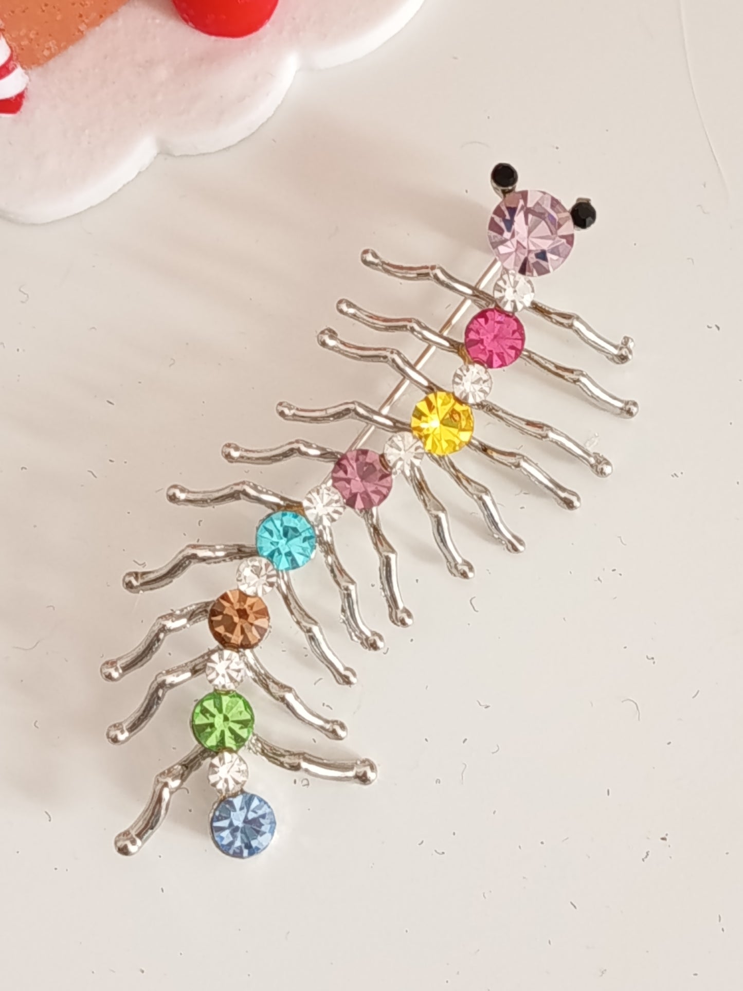 Une broche fantaisie en forme de mille-pattes argenté sertie de strass colorés. La broche est posée sur une sur une surface blanche à côté d'une petite maison miniature.