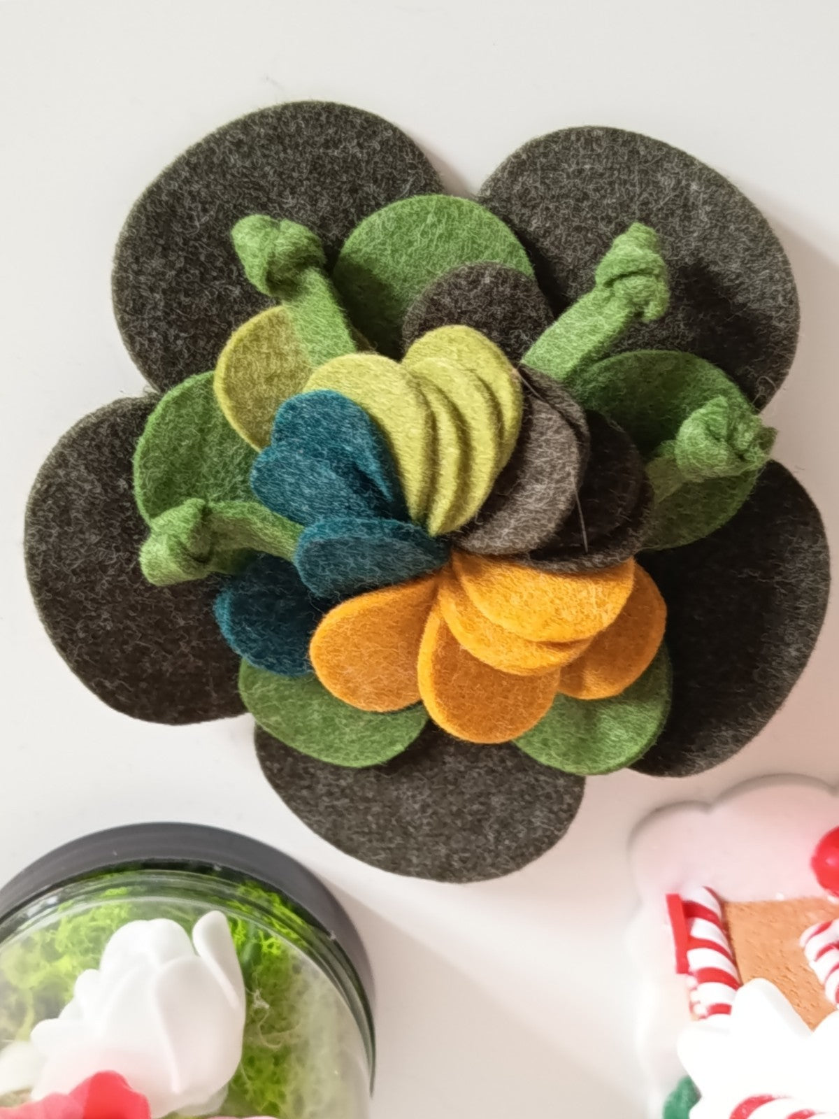 Broche artisanale en feutrine de laine,  en forme de fleur multicouches, coloris vert forêt, olive, jaune moutarde sur fond blanc.
