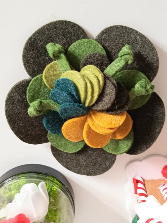 Broche artisanale en feutrine de laine,  en forme de fleur multicouches, coloris vert forêt, olive, jaune moutarde sur fond blanc.
